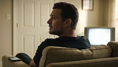 Man sitting on beige sofa turning left with smartphone on armrest and tv glowing
