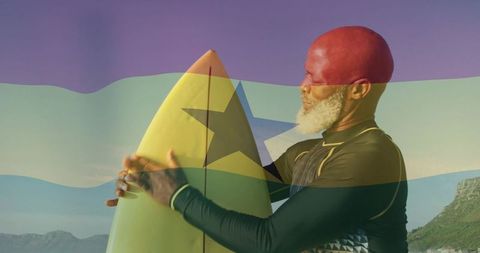 Mature man cradling yellow surfboard with black star and flag overlay, coastal sunset