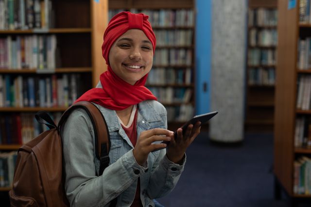 Asian female student wearing a red hijab is studying in a library, using a tablet computer. She is sitting between bookshelves, smiling, and appears engaged in her work. This image can be used for educational content, technology in education, diversity in learning environments, and promoting digital learning tools.