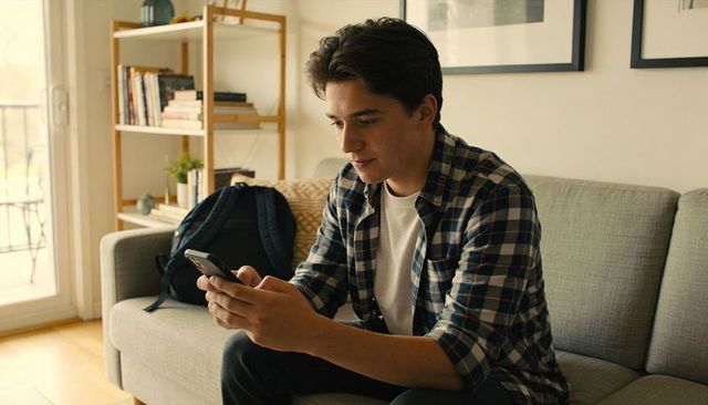 Young man in plaid shirt sitting on sofa texting on smartphone in bright modern apartment