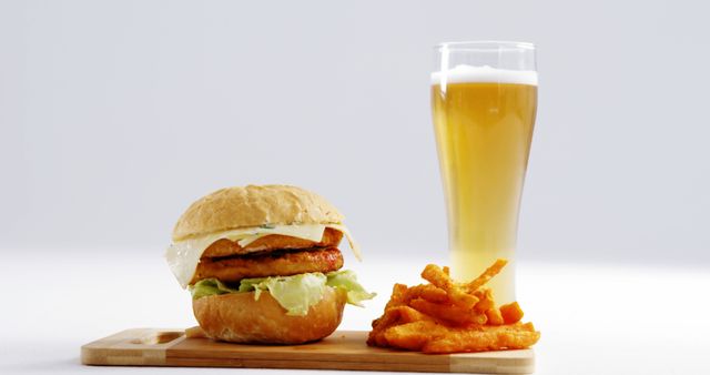 This stock photo shows a gourmet chicken burger with lettuce and a light sauce, accompanied by crispy fries and a cold beer served in a tall glass. Ideal for use in advertising for pubs, restaurants, or food delivery services. This image also works well for blog posts, social media marketing, and menu designs focusing on casual dining and fast food.