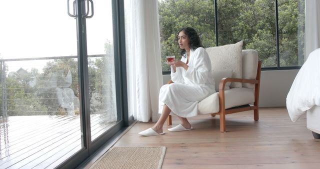 Woman enjoying a peaceful moment drinking tea in a modern, light-filled bedroom with large windows offering a scenic nature view. Ideal for themes related to relaxation, serene mornings, self-care, and comfortable living.