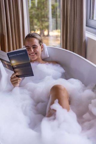 Smiling woman relaxing in freestanding tub with bubbles reading endless sunsets paperback