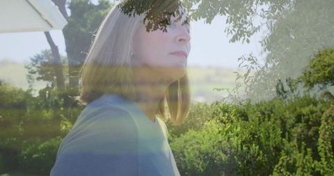 Standing mature woman gazing upward in sunlit garden, contemplative portrait with lens flare