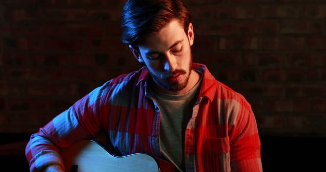 Man wearing a red plaid shirt playing an acoustic guitar indoors against a brick background under ambient lighting. Ideal for themes related to music, solo performance, musicians, hobbies, or creative expression.