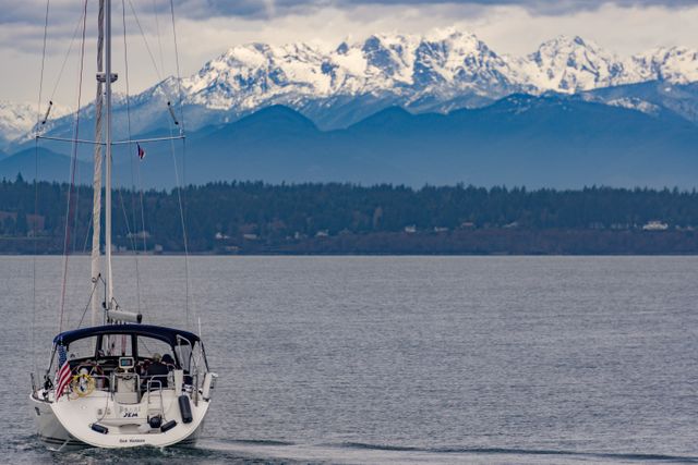 Sailboat gliding on calm water with snow-capped mountains looming in the background, creating a peaceful and scenic view. Ideal for use in travel blogs, adventure and outdoor content, marketing materials for travel agencies, and website headers promoting relaxation and nature retreats. This stock photo showcases an exceptional landscape combining nautical and mountainous elements, emphasizing tranquility and exploration.