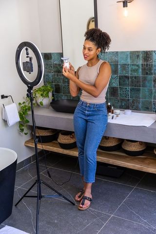 African american woman filming skincare tutorial with ring light and smartphone in bathroom