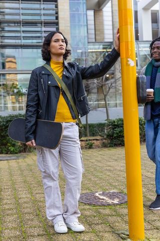 Skateboarder leaning on yellow pole with friend holding coffee in city plaza