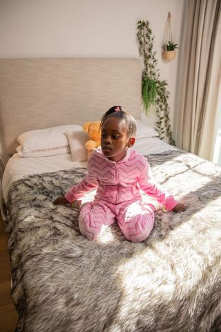 African american preschool girl sitting on cozy bed wearing pink onesie with sunlight and teddy