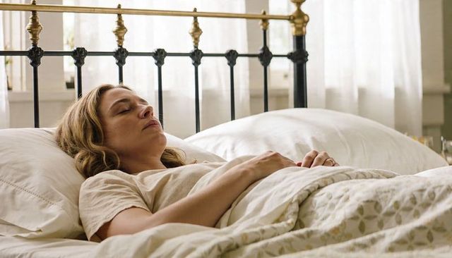 Middle-aged woman resting peacefully on white linens in sunlit bedroom with metal headboard