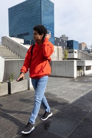 African american teen walking in red hoodie adjusting headset on wet urban plaza