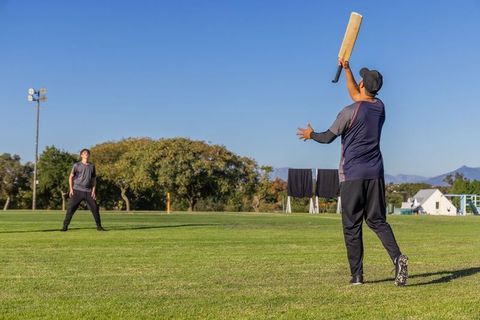 Male teammates practicing cricket batting with wooden bat and teenage fielder on sunny outfield