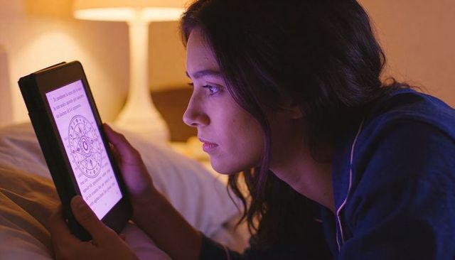 Woman reading e-reader in bed at night, holding device close in piped pajamas