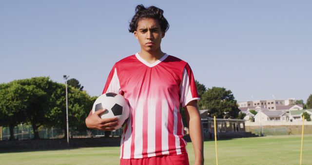 Teen athlete wearing a red and white striped soccer uniform holding a soccer ball on an outdoor field. Trees and buildings visible in the background. Useful for sports-related promotions, youth athletic programs, confidence campaigns, and team spirit visuals.