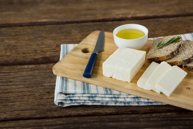 Gouda cheese and brown bread slices arranged on a wooden chopping board with a small bowl of olive oil and a knife. Ideal for use in food blogs, recipe websites, healthy eating promotions, and culinary magazines.