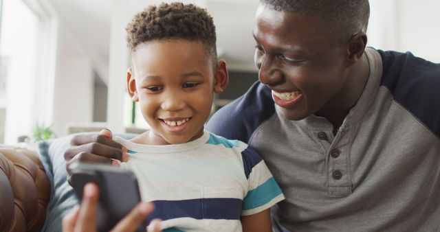 Image of happy african american father and son sitting on sofa and using smartphone. Family, spending quality time together at home with technology concept.