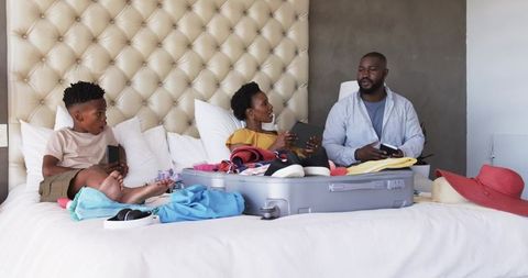 African American family packing suitcase on bed for weekend getaway using smartphones