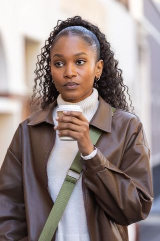 Young black woman sipping takeaway coffee while walking city street wearing leather jacket