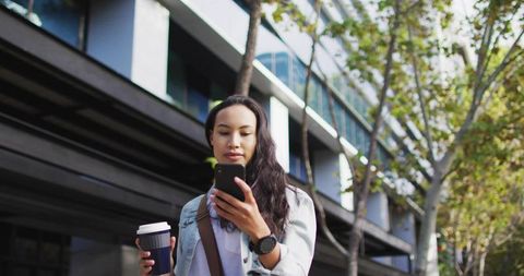 Walking woman carrying coffee checking smartphone by modern office facade