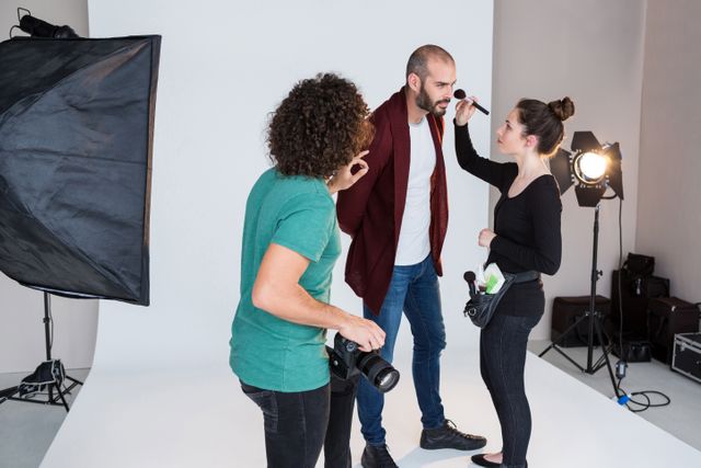 Makeup artist applying makeup to model in a professional studio setting. Photographer and lighting equipment visible, indicating a collaborative effort in a fashion or beauty photoshoot. Ideal for use in articles about the fashion industry, beauty tips, photography techniques, or behind-the-scenes content.