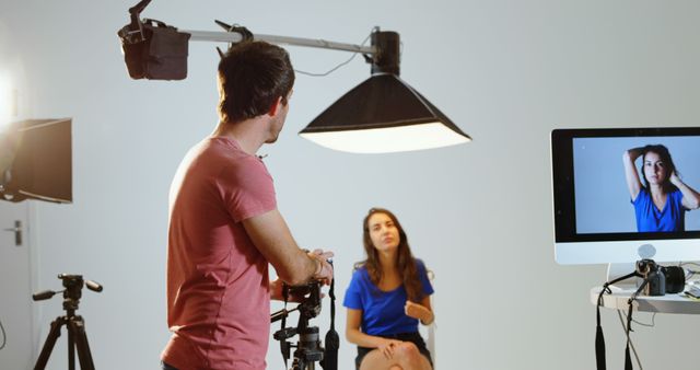 This image depicts a behind-the-scenes view of a photography studio. A female model is posing in front of a camera while a professional photographer is adjusting equipment. The scene shows lighting setups and a computer monitor displaying the live shot. Ideal for topics related to professional photography, media production, or advertisement materials highlighting photography services.