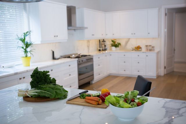 Fresh vegetables including carrots, bell peppers, and leafy greens are on a marble kitchen worktop. The modern kitchen features white cabinets and a clean, bright atmosphere. Ideal for use in articles or advertisements about healthy eating, home cooking, kitchen design, and lifestyle.