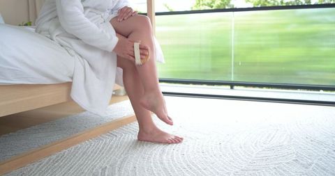 African american woman dry brushing leg on low wooden bed in sunlit bedroom for self care