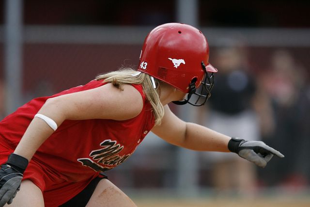 Female softball player wearing a red uniform and helmet, preparing to field the ball. Great for use in sports-related content, showing athletic focus and determination or promoting women's softball events and equipment.