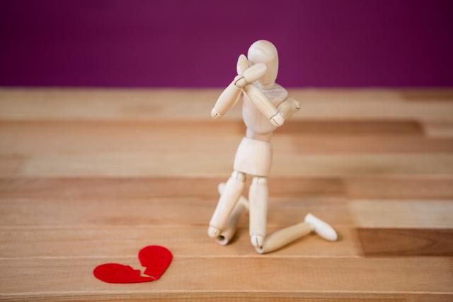 Wooden figure kneeling with hands covering face beside broken red heart, conveying emotions of sadness and heartbreak. Useful for illustrating themes of relationships, emotional struggles, breakups, emotional healing, and psychological concepts in articles, blogs, and counseling materials.
