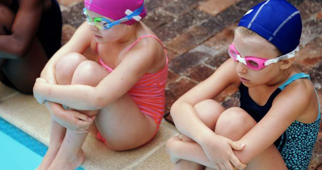 Two children in colorful swimwear and caps are sitting by the edge of a pool wrapped in their arms. They are wearing goggles and seem focused. This image can be used for topics related to swimming lessons, recreational activities, childhood, sports programs, or family outings.