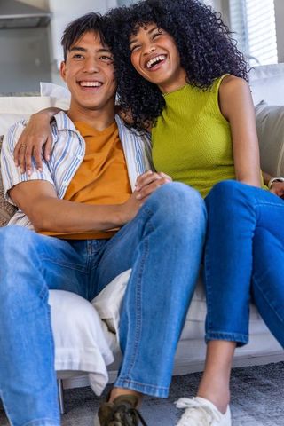 Laughing asian man and african american woman cuddling on sofa at home in casual clothes
