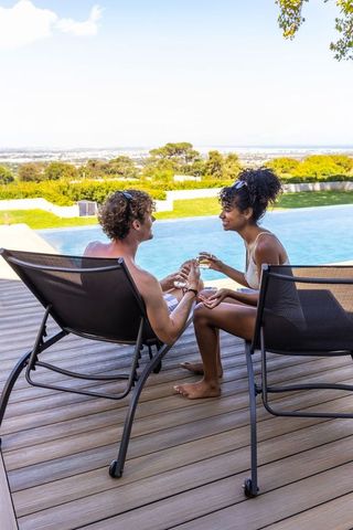 Diverse couple relaxing by private pool on sunlit deck sharing drinks and foot massage