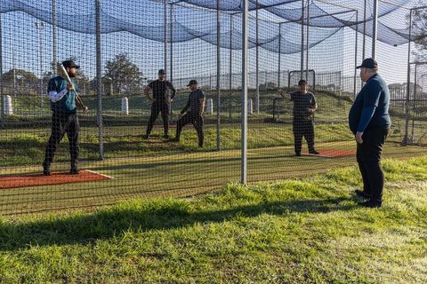 Baseball coach leading batting cage drill with batter swinging wooden bat and trainers