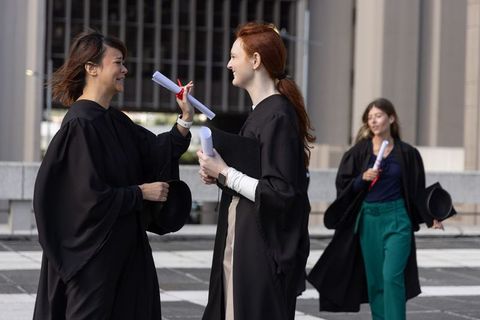 Female graduates celebrating on campus plaza holding diplomas in black gowns and mortarboards