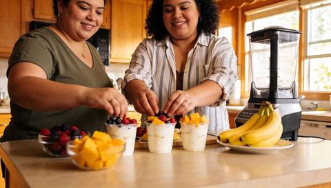 Two women preparing yogurt parfaits with fresh berries and mango in sunny home kitchen