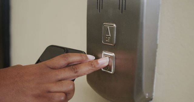 Close-up view of person's hand pressing an elevator button while holding a smartphone, showcasing urban life and modern technology. Ideal for illustrating convenience, daily routines, and technological integration in advertising, educational materials, or articles about urban living.