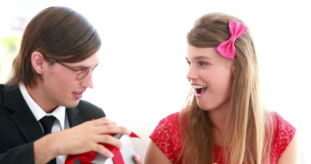 Young woman in red dress with pink hair bow expressing excitement as her boyfriend presents a gift. Ideal for illustrating concepts of romance, surprises, special occasions, and loving relationships.