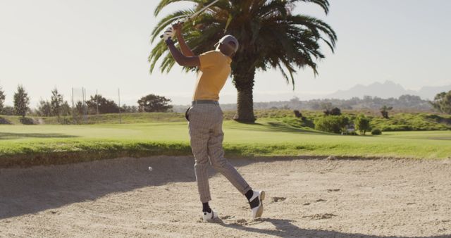 Male golfer in action hitting a shot from a sand trap on a lush golf course in sunny weather, surrounded by palm trees. The image is perfect for promoting sporting events, advertising golf gear, or illustrating articles about golfing techniques.