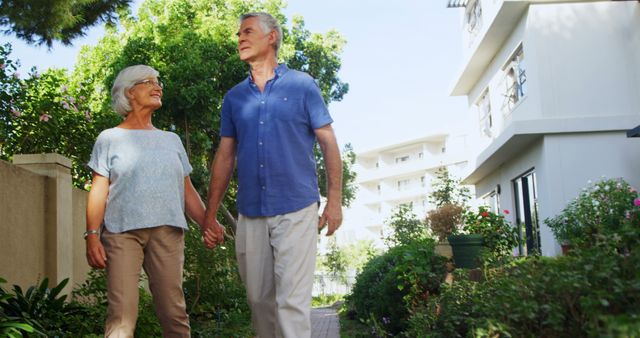 Senior couple walking hand in hand in an urban garden setting. They are smiling and looking at each other, surrounded by lush greenery and residential buildings. Great for themes related to retirement, active aging, companionship, healthy lifestyle, urban living.