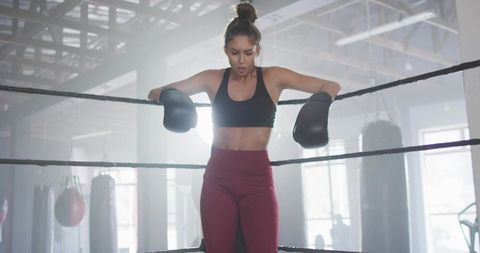 Woman boxer leaning on ropes after intense training in industrial gym backlit by windows