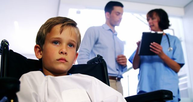 Boy in wheelchair appears concerned while doctor and parent discuss in background. Suitable for themes around pediatric healthcare, medical treatment, parenting, hospital environment, patient care, healthcare services, or medical consultations.