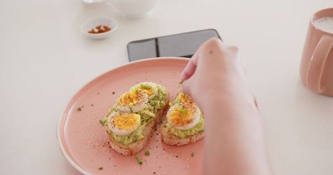 Avocado Toast Being Seasoned on Coral Plate with Smart Device Nearby