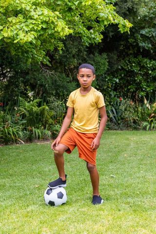 African american boy standing with soccer ball on sunlit backyard lawn, summer play