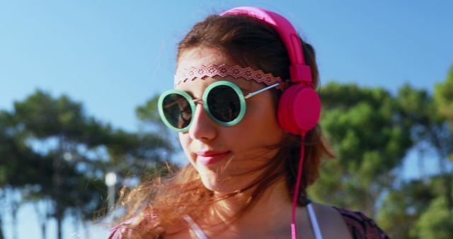 Young woman listening to music with pink headphones and round sunglasses, standing outdoors on a sunny day. Ideal for use in travel blogs, music promotions, fashion posts, or lifestyle content showcasing carefree and joyful moments.