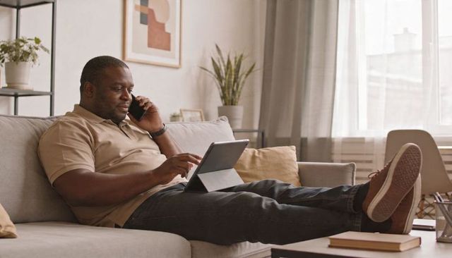 Reclining man on sofa using tablet while talking on phone in cozy modern living room