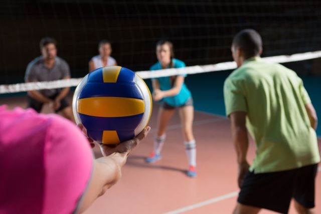 Female volleyball player preparing to serve ball during match with teammates in background. Ideal for sports articles, team spirit promotions, athletic training materials, and competitive sports advertisements.