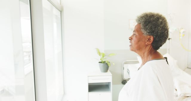 Elderly woman stands by large hospital window in brightly lit room, lost in thought. Ideal for use in stories or articles about aging, healthcare, reflection, medical conditions, or emotional moments in healthcare settings.