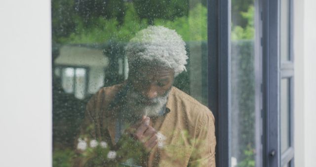 Senior man gazes thoughtfully out a rain-covered window. This could be used in content related to aging, contemplation, mental health, solitude, or rainy day reflections. Ideal for articles or advertisements centered on introspection, loneliness, or serenity.