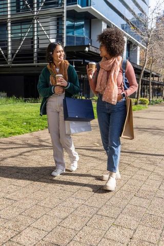 Multicultural women walking with coffee and shopping bags on sunlit urban sidewalk