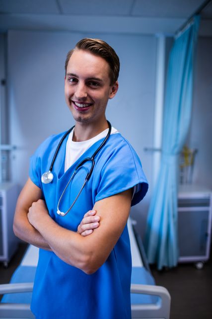 This image shows a confident male doctor standing with arms crossed in a hospital ward, smiling warmly. He is wearing blue scrubs and a stethoscope around his neck, indicating his role as a healthcare professional. This image can be used for medical websites, healthcare promotional materials, hospital brochures, and articles related to patient care and medical staff.